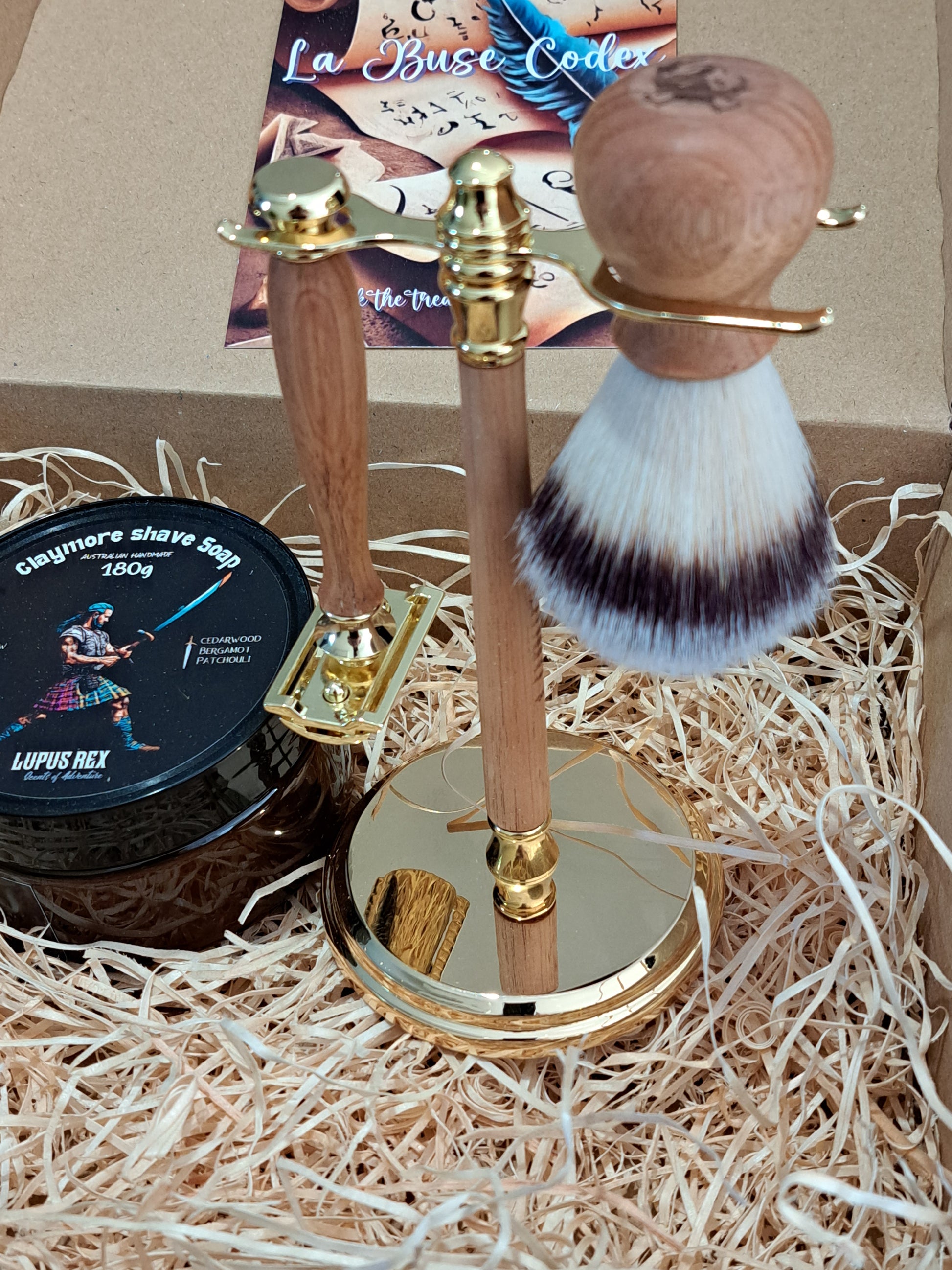 Timber stand with gold coloured metal fittings with the synthetic knot timber shave brush and timber handle gold coloured safety razor and claymore tallow based shave soap in 100% recycled cardboard La Buse gift box